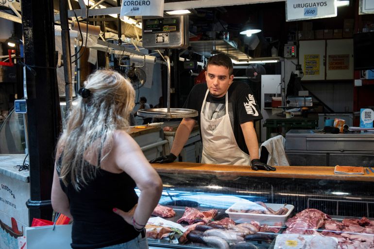 A woman stands in front of a meat counter, as a butcher waits to take her order