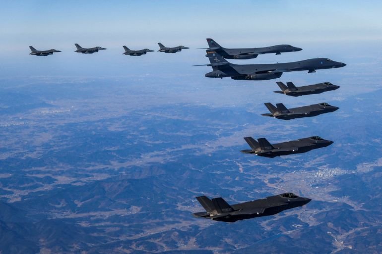 US Air Force B-1B bombers and F-16 fighter jets along with South Korean Air Force F-35A fighter jets take part in a joint air drill on February 19, 2023. They are in a V-shaped formation in the sky over land.