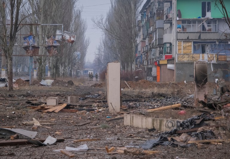 The ruined city of Bakhmut. There are trees along the road and an apartment block damaged by shelling. There are piles of debris on the ground and road. A few Ukrainian soldiers are in the background.
