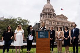 Amanda Zurawski speaks in front of the Texas State Capitol. Three women stand to her left on the grass and three to her right, with a male next to them. Amanda stands behind a pulpit that has a sign on it reading, "Center for Reproductive Rights".