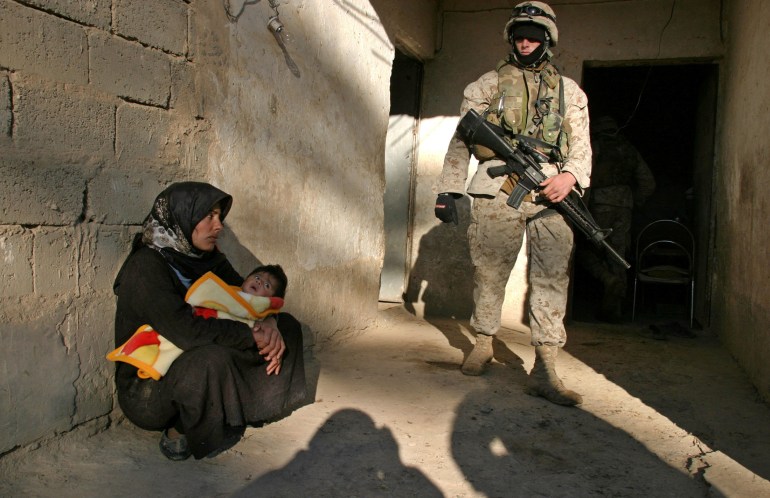 A woman lies crouched on the ground with a child in her arms, while a US army soldier stands by