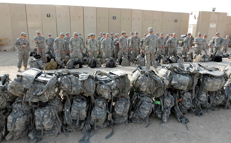 Two rows of backpacks lie in front of lines of soldiers and the back of a commander talking to them. They are dressed in grey and white camouflage and the bags are in the same pattern.
