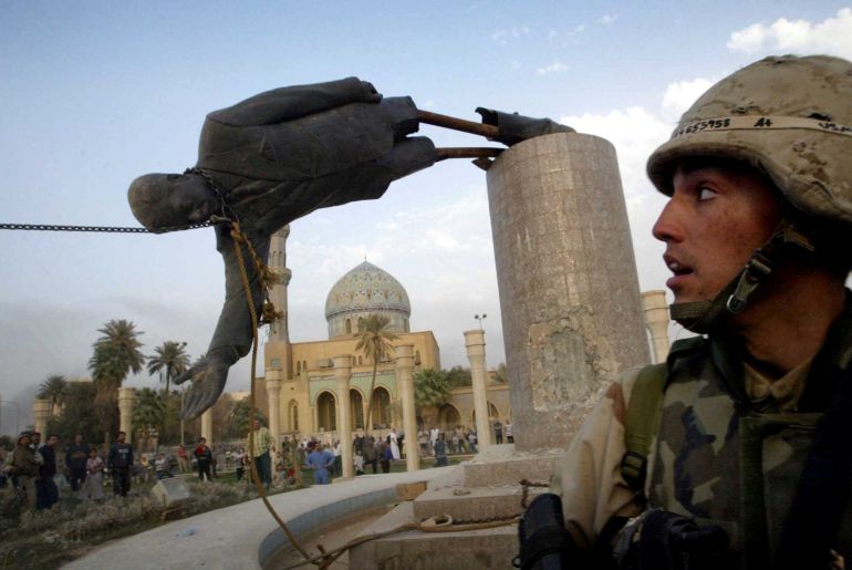 A man in the foreground watches as a giant statue falls in the center of Baghdad