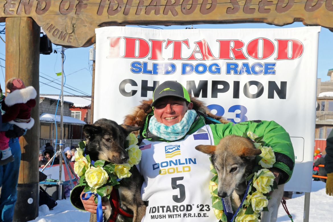 A man holds two dogs, one under each arm, in front of a sign that reads "Iditarod sled dog race champion"