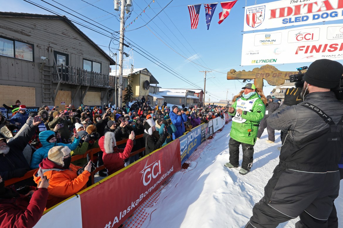 A man in a green winter coat greets fans at the Iditarod finish line
