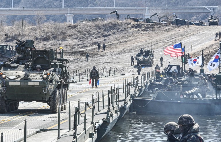 A personnel carrier on a temporary bridge with some small boats alongside. There are soldiers walking in front and behind, as well as some on the boats, which are flying US and South Korean flags. 