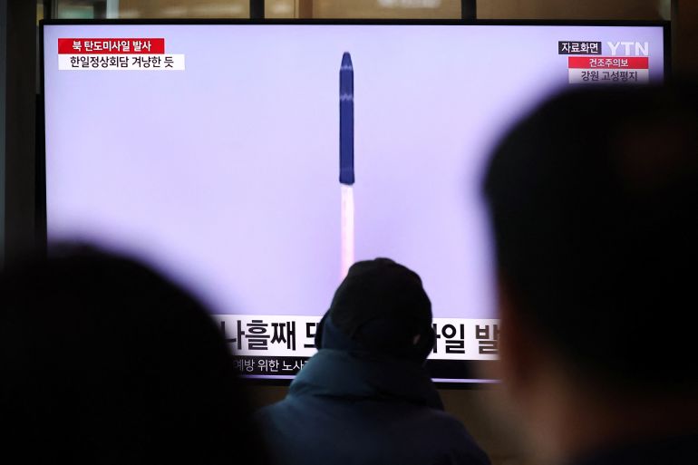 A large screen at a railway station in Seoul, South Korea, shows a TV news report on North Korea firing a ballistic missile. A few people's heads are silhouetted against the bottom of the screen, giving the impression that they are watching the broadcast.