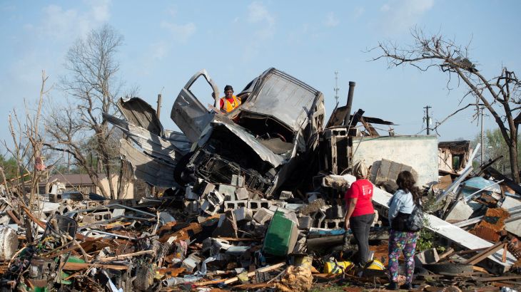 Residents of Rolling Fork, Mississippi, survey damage from a tornado. A visibly-damaged car, possibly a SUV, is lying on top of a mound of debris.