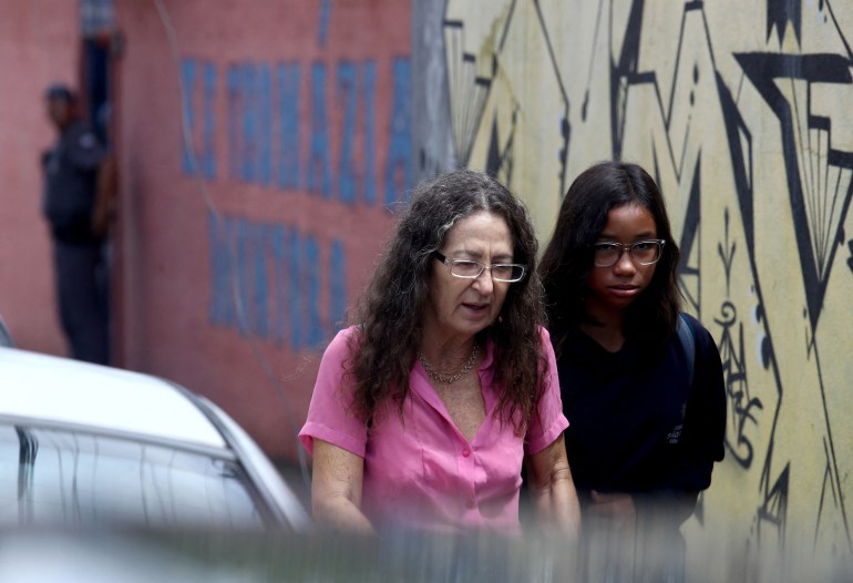 A woman and a teenaged girl walk between a wall and a car, heads bent.