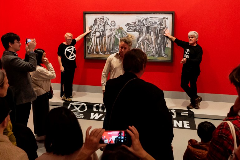 Two extinction rebellion protesters glued to a Picasso artwork at the National Gallery of Victoria in Melbourne, Australia. They are dressed in black and the walls are red. There is a crowd of onlookers.