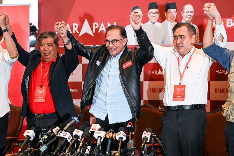 Ibrahim wearing a black leather jacket stands in the middle of members of his team. They are in a row and holding each others' hands raised over their heads. There are many microphones pointed at them.