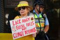 A woman holding a sign in support of Deanna 'Violet' CoCo. It reads 'Lock away fossil fuels not climate defenders'. The woman is wearing sunglasses and a straw hat with yellow flowers around the brim. There is a policeman in the background.