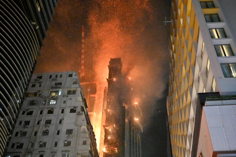 A 42 storey high rise building on fire in Kowloon, Hong Kong. There are orange flames, embers floating in the night sky and smoke. There are other skyscrapers nearby
