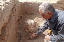 A man brushes sand away from the unearthed head of a smiling sphix statue.