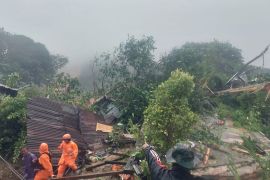 Landslide in the Natuna region. There some rescuers in front in bright orange suits and lots of mud and debris behind them. It is still raining.