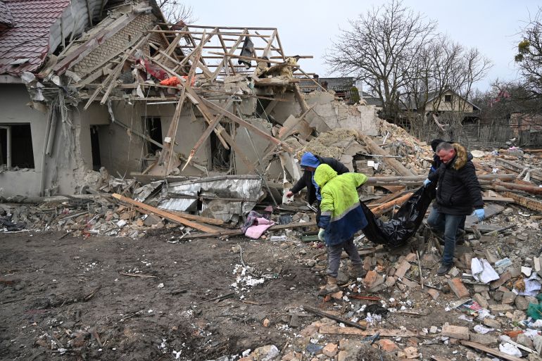 Police and local residents at the site of a Russian missile attack. There are piles of rubble. They are carrying a body bag out.