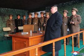 Kim Jong Un in a black coat standing next to a wooden desk in a shelter. Behind him are a row of military officers in uniform.