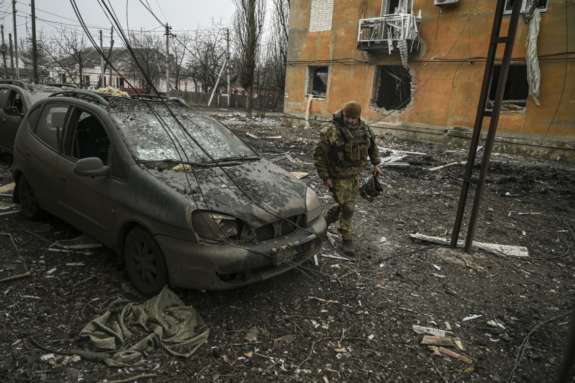 A Ukrainian service member looks at a burned-out car after shelling in the village of Chasiv Yar, Ukraine. The street around him is littered with debris and the building in the back seems hollowed out with all its windows blown out.