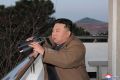 Kim Jong Un in a brown jacket holding binoculars as he leans against a balcony railing.