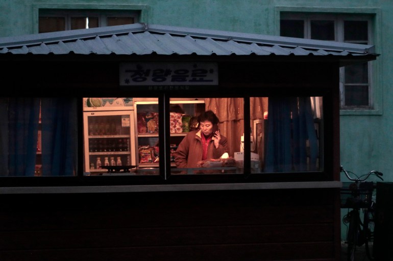 It appears to be night time, but the rectangle of light behind the booth window shows a lady in a brown jacket talking on the phone.  Behind it is a drinks fridge.