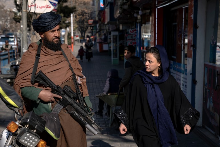 A Taliban guard stands guard as a woman walks past in Kabul, Afghanistan, on December 26, 2022 [Ebrahim Noroozi / AP Photo]