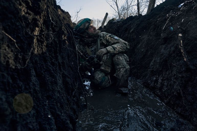 A Ukrainian soldier crouches down in a trench near the front line in Bakhmut, Ukraine. He's looking to the sky and has his weapon in his right hand. It's very muddy.