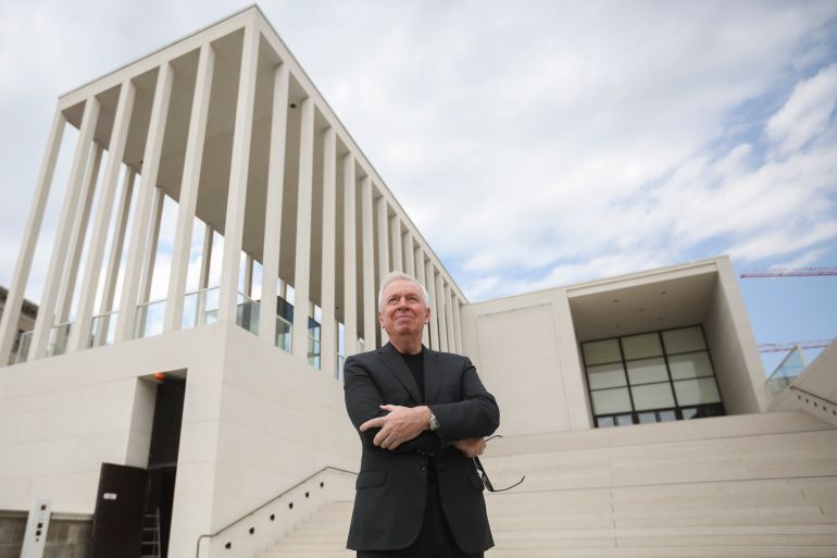 David Chipperfield outside the James-Simon-Galerie at Berlin's Museums Island in Germany. He is standing with his arms crossed at the bottom of some white steps. The building is in an elevated position behind him. It is white and flat-roofed with slender white columns.