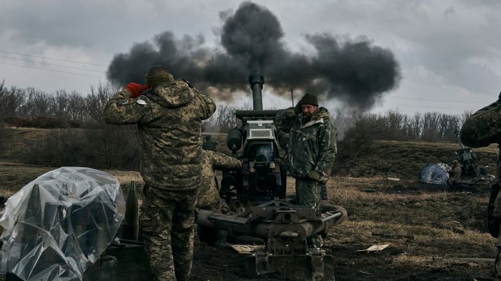 Ukrainian soldiers cover their ears after firing a howitzer. There is black smoke coming from the howitzer which is facing towards trees and brownish grass.