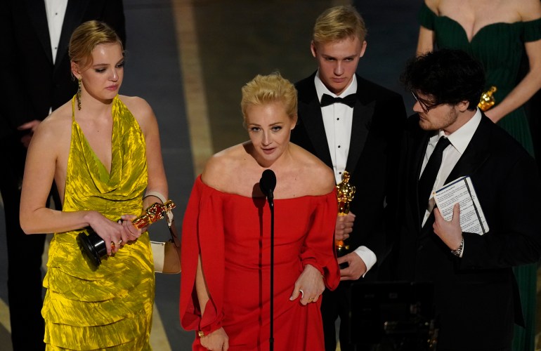 Kırmızı elbiseli bir kadın, film yapımcılarıyla çevrili bir mikrofona konuşuyor.