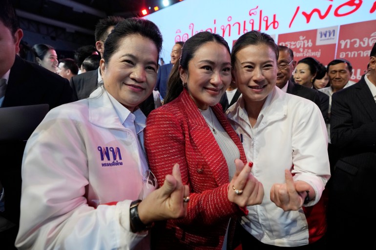 Paetongtarn Shinawatra with two supporters at a Pheu Thai rally. She's wearing a red jacket and is smiling. They are making the mini heart gesture with their fingers.