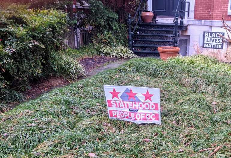 DC statehood yard sign. It is small and white, pinned into the grass. There are three pink stars at the top and below the words: STATEHOOD, PEOPLE OF DC 