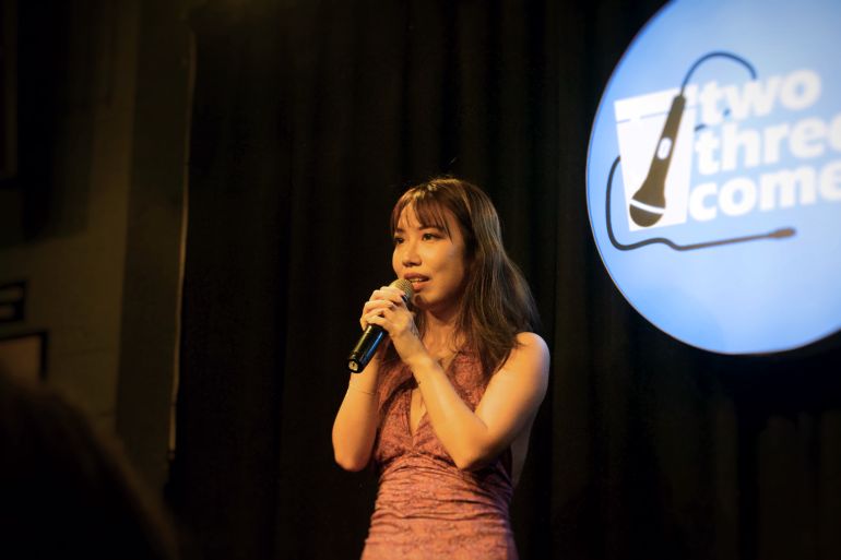 Jamie Wang performing at a comedy night. She's wearing a pink sleeveless dress and speaking into a microphone. Behind her the stage is black except for a blue circle with the words Two Three Comedy printed in white and a drawing of a microphone