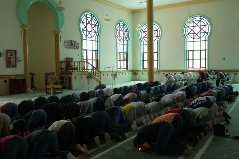 The inside of the mosque is dark but tinged with a green light coming through the greenly framed windows. Worshippers are in rows in the middle of prayer, on their knees, with heads bowed to the ground. There is a small fan at the front that seems too small for the large area it is cooling.
