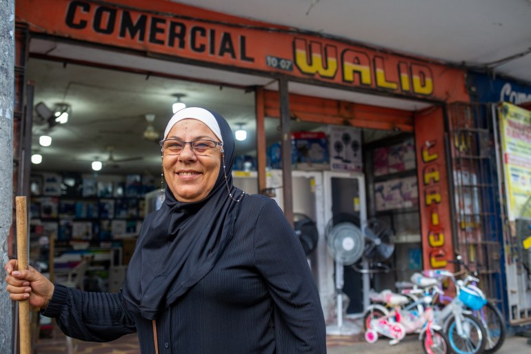 Samira Hajj Ahmad holds a broom outside her electronics shop. She is smiling and wearing a dark hijab. Behind her, the shop has a kids' bicycles standing out front and a couple of fans. Above the shop are the letters: 'COMMERCIAL WALID'