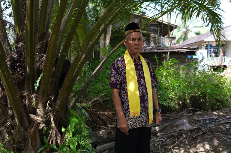 Sibukdin standing near an oil palm tree with houses behind him.  He is wearing a patterned shirt and with a yellow scarf with tassels at the end draped around his neck.  He is also wearing a traditional black hat with gold embroidery.