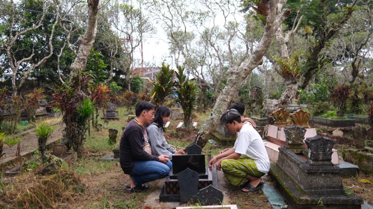 Wiyanto, his daughter and Septian’s friends pray together by his grave, which is near their family home. They are kneeling down. Wiyanto is on the left of the grave. There are gnarled trees around the cemetery.