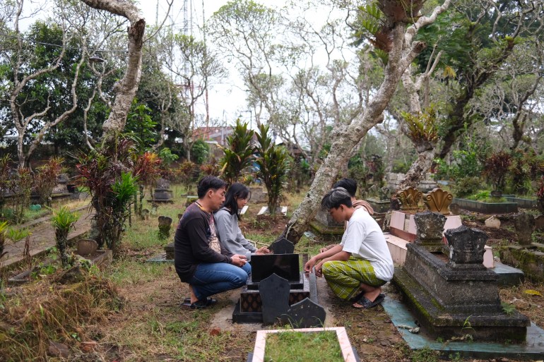 Wiyanto, his daughter and Septian’s friends pray together by his grave, which is near their family home. They are squatting down. Wiyanto is on the left of the grave with his daughter. The friends are opposite them. There are gnarled trees around the cemetery.
