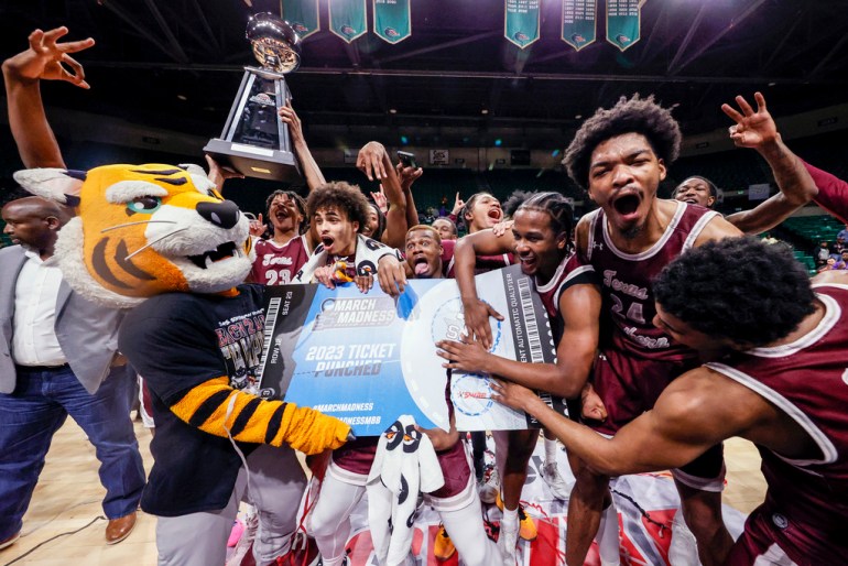 Texas Southern basketball players celebrate after beating Grambling State to earn their ticket to March Madness.  A team mascot holds a large sign saying "2023 STAMPED TICKET" as players gather around her.  A player in the background holds a trophy.