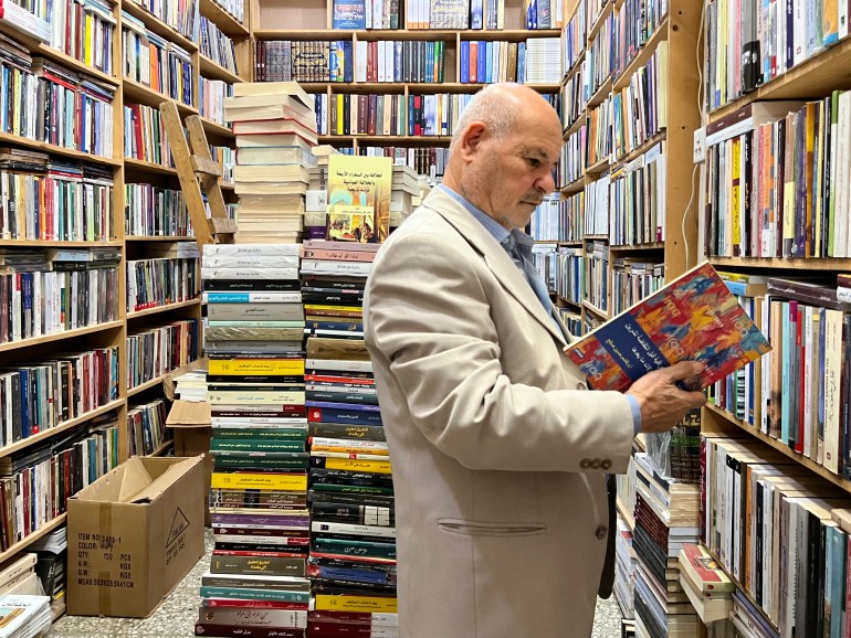 A man looks through a book in a room surrounded on three sides with bookshelves, and more stacks of books behind him. 