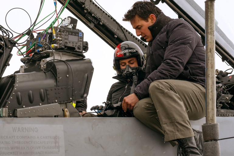 Tom Cruise sits on the edge of a fighter plane, as Monica Barbaro sits inside