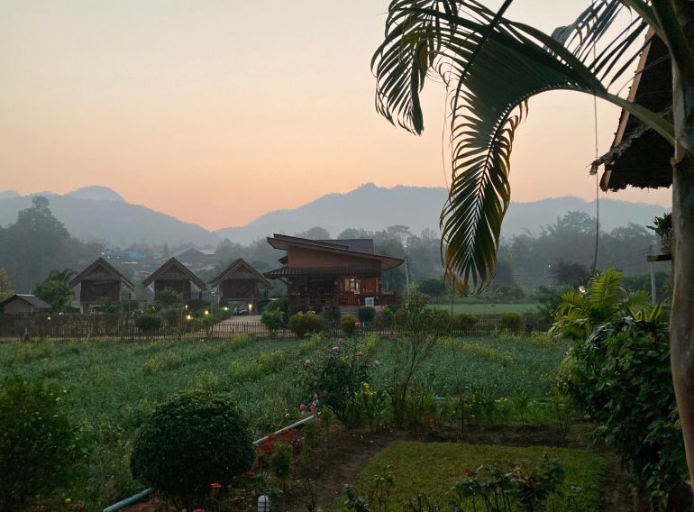 An intense red sunset behind mountains in northern Thailand. There are wooden buildings set among lush green fields in front and palm fronds to the right.