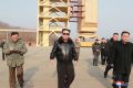 Kim Jong Un walks with officials at the Sohae Satellite launch site. He is wearing a black leather jacket, baggy black trousers and sunglasses. A launch structure is behind him