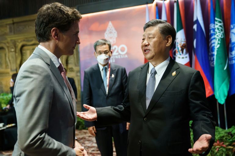 Xi Jinping speaks, with his arms outstretched, as Justin Trudeau listens in a conference room. A man stands behind them both wearing a mask.