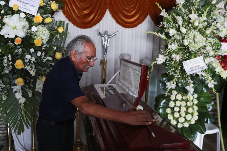 A man places a white rose atop a casket, surrounded by flowers