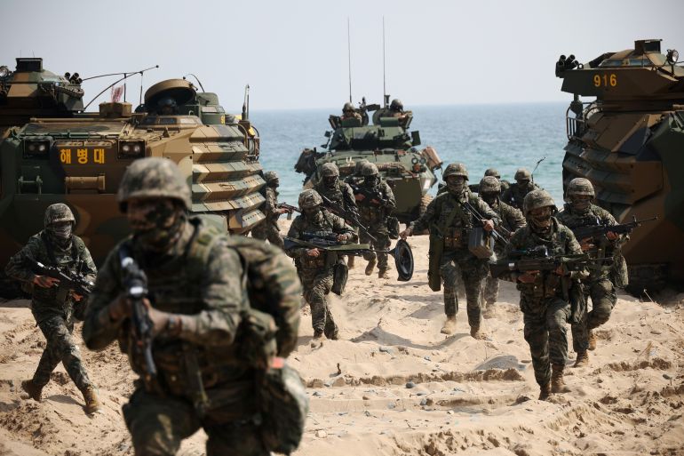US and South Korean soldiers running up a beach during military drills. There are military vehicles on either side of them and behind them. They have their weapons ready.