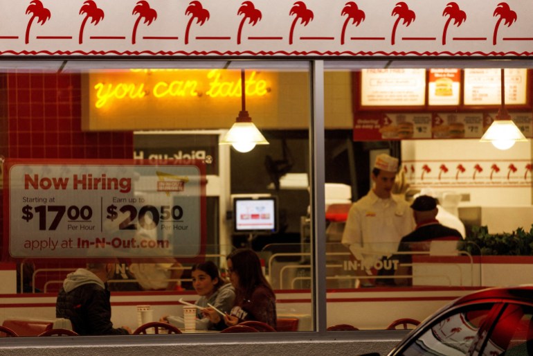 A "Now hiring" sign is displayed on the window of an IN-N-OUT fast food restaurant i