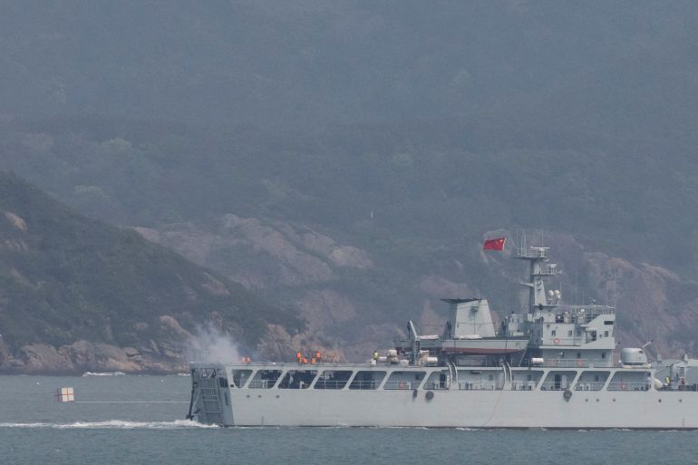 A Chinese warship fires its gun. The weapon is at the rear of the vessel, which is flying the Chinese flag. There are mountains behind.
