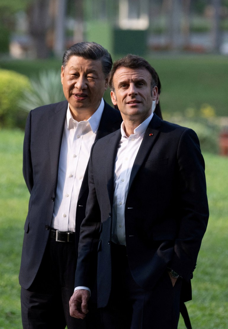Xi and Macron walking in a garden in southern China. Both men are wearing dark suits and open-necked white shirts. They look relaxed.
