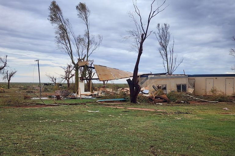 The ruined Pardoo Roadhouse after Cyclone Ilsa. The roof has been torn off, and debris is lying around. The trees have been shredded. The sky is heavy with cloud