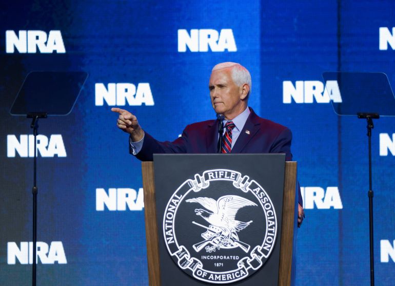 Mike Pence speaking at the NRA convention. The NRA logo is repeated across the blue backdrop behind him. He is pointing out into the audience and looking serious.